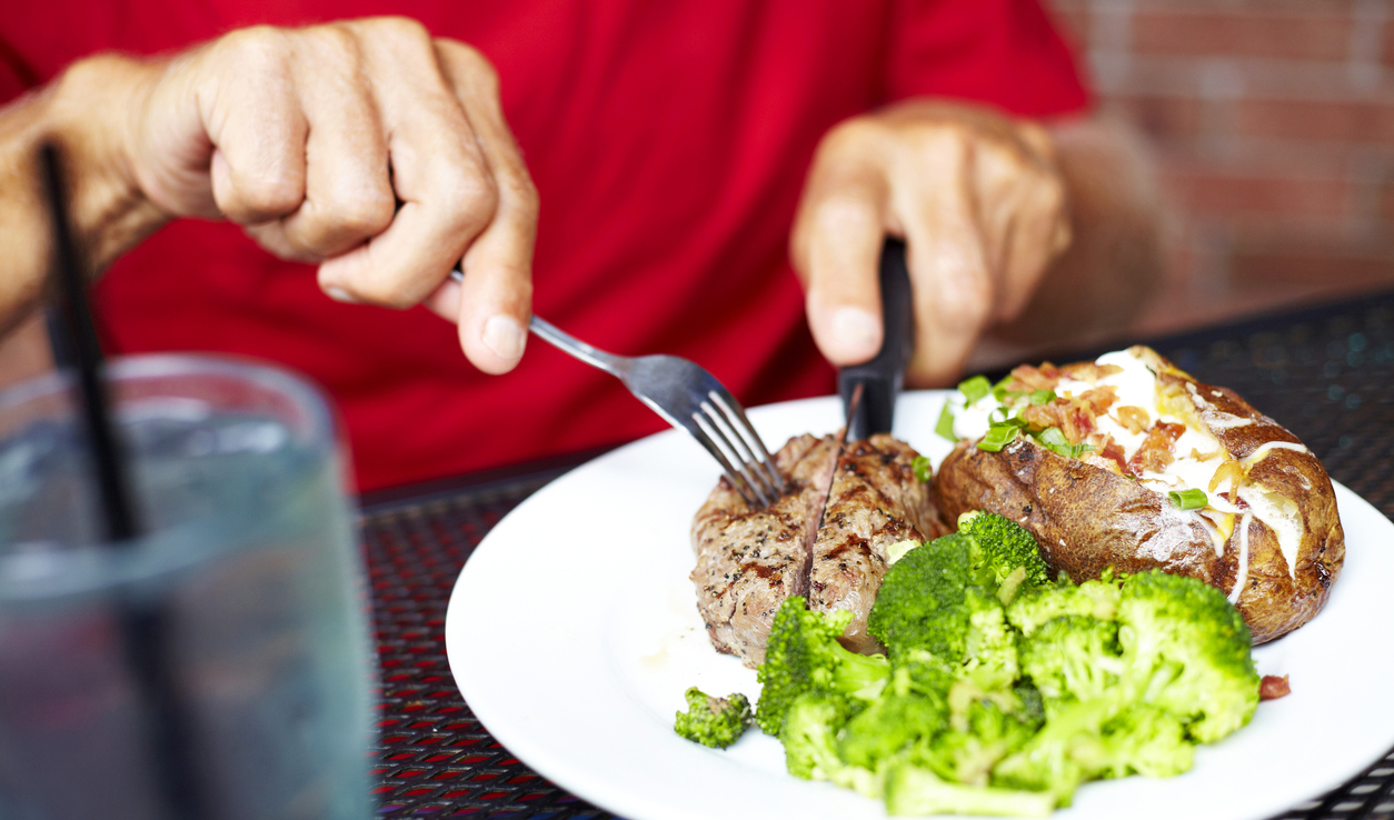Midsection of senior man eating strip steak served with loaded baked potato and broccoli at restaurant table