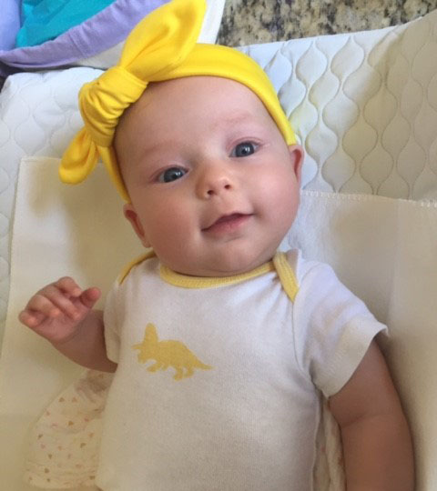 Baby girl with yellow bow on her head with white onesie and single yellow dinosaur print