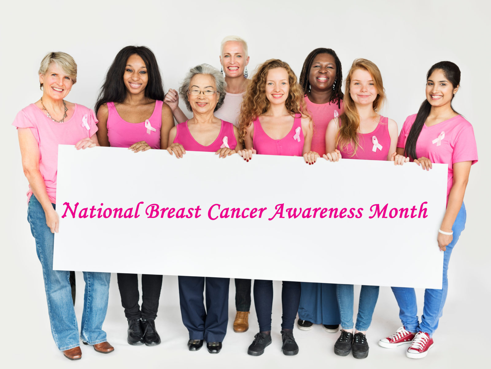 Group of women of various ages wearing different shades of pink shirts, holding National Breast Cancer Awareness Month sign.