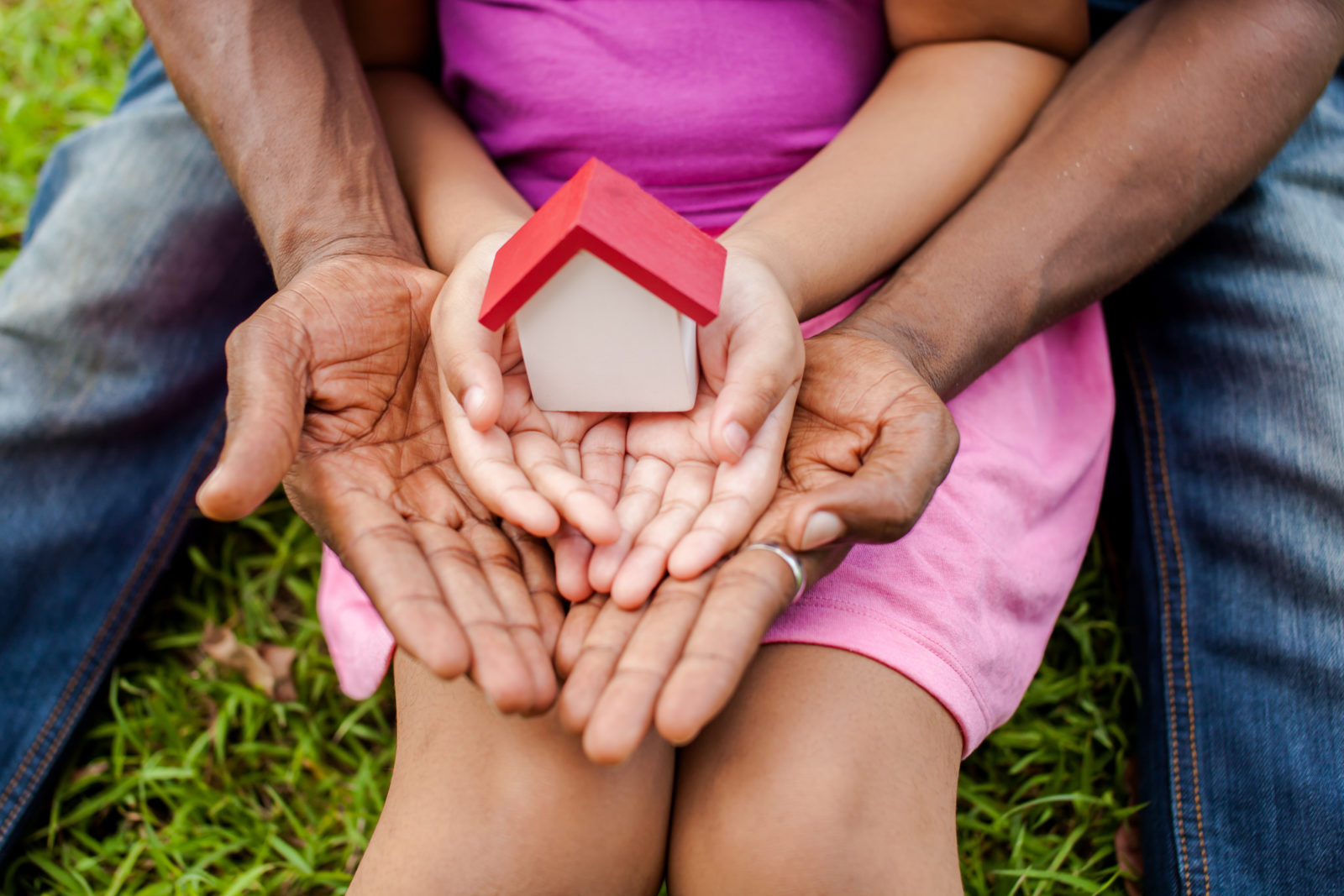 African American father sitting on grassy ground with legs spread apart. Little African American girl sitting in front of him (with her back against him) holding a wooden house in her opened palms with father's hands underneath her's.