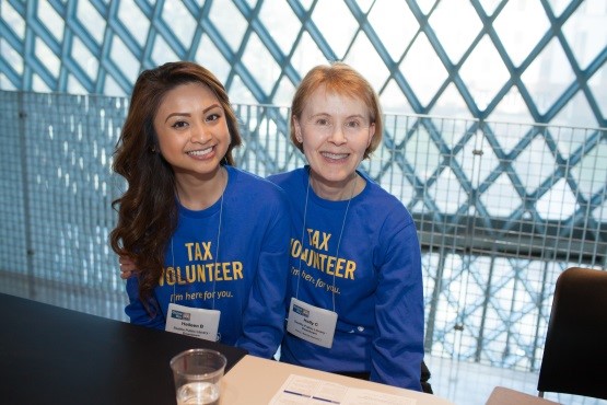 Two smiling volunteers wearing blue Tax Volunteer t-shirts are sitting at a table.