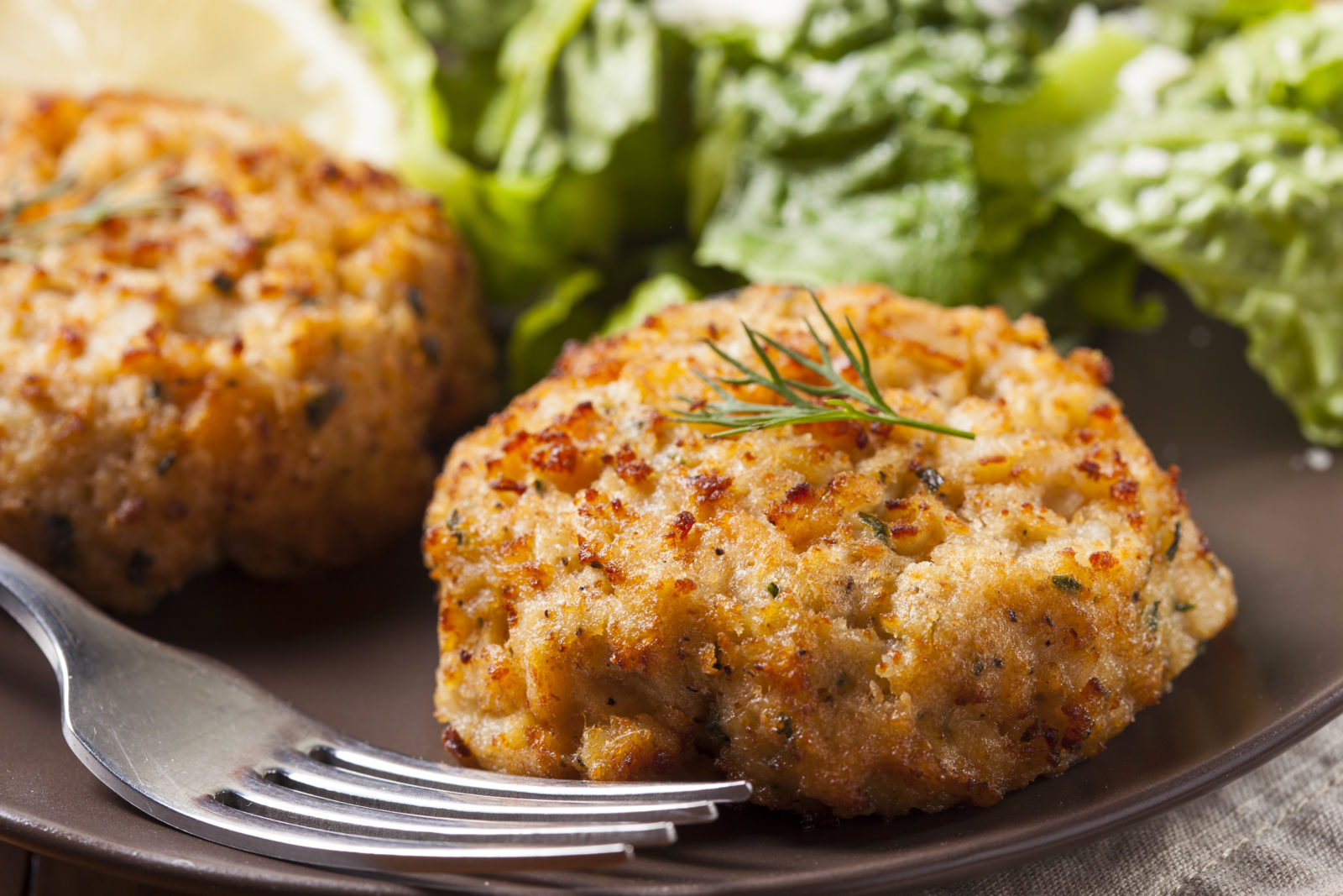 Two delicious looking fish cakes on a plate with a fork in the foreground and salad in the background of the plate.