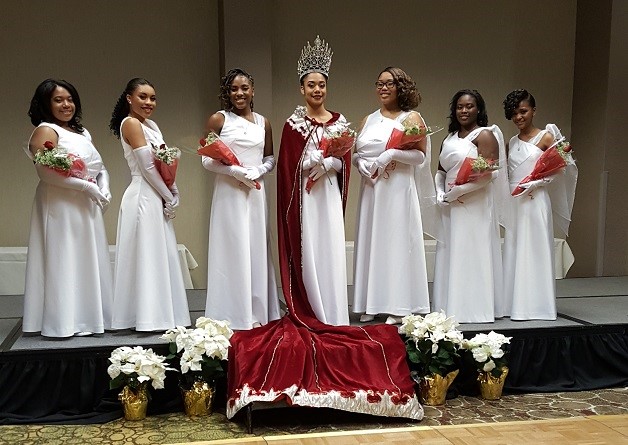 Seven young African American women posing in a court formation with the Queen in the Center wearing a rhinestone crown with a long red regal cape draped on the floor.