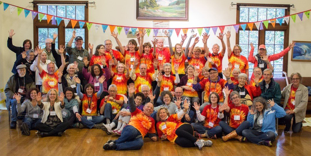 Colorful photo of a crowd of people wearing red, orange, and yellow tye-dyed tshirts, waving at the camera. Some are standing, with a banner of pennant flags behind them. Some are seated. Some are stretched out on the floor in happy, relaxed poses.
