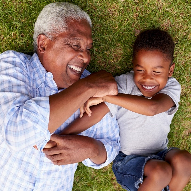 Photo of African American grandfather and grandson, laughing and wrestling on a green lawn.