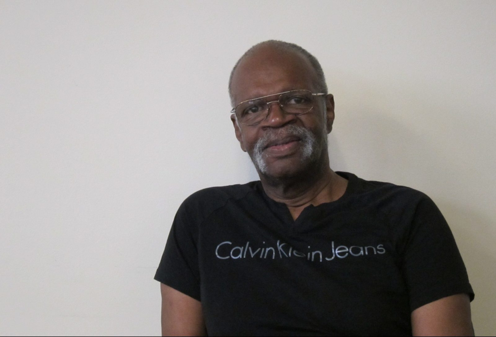 photo of Mr. Greg Francis, who is the subject of this article. Mr. Francis is an older African American man with close cropped hair, eye glasses, and a gray moustache. He is wearing a black t-shirt with what appears to be a Calvin Klein Jeans logo across the chest.