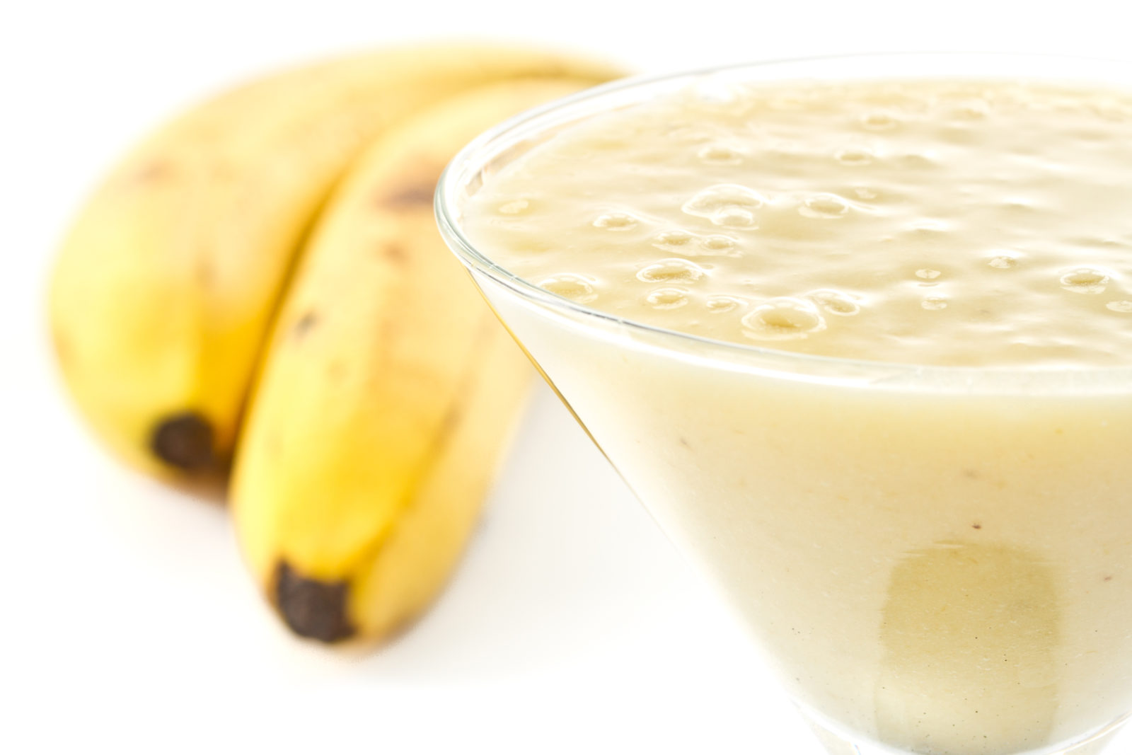 Photo of two ripe yellow bananas and a glass of bubbly liquid smoothie on a white surface.