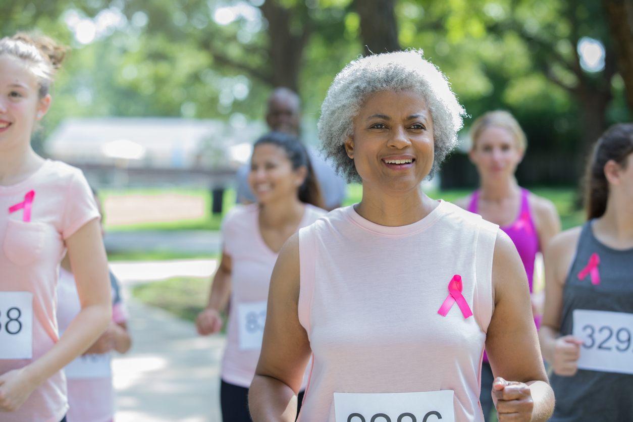 Beautiful senior adult African American woman is smiling while running in race to benefit the cure for breast cancer. She is wearing pink athletic clothing and a pink ribbon. Teammates are running behind her.