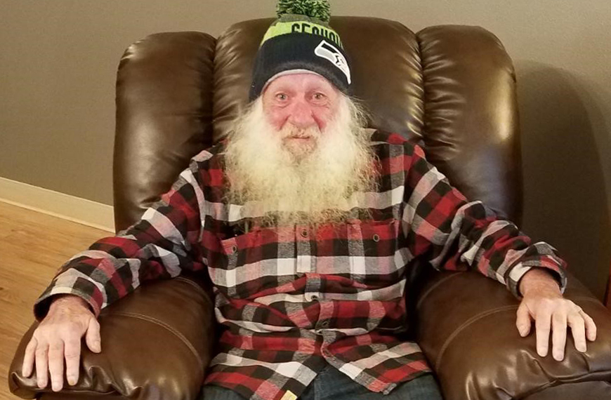 photo of Larry Stuvland, an older man with a long white beard, wearing a plaid shirt and Seahawks hat and sitting in a large leather armchair