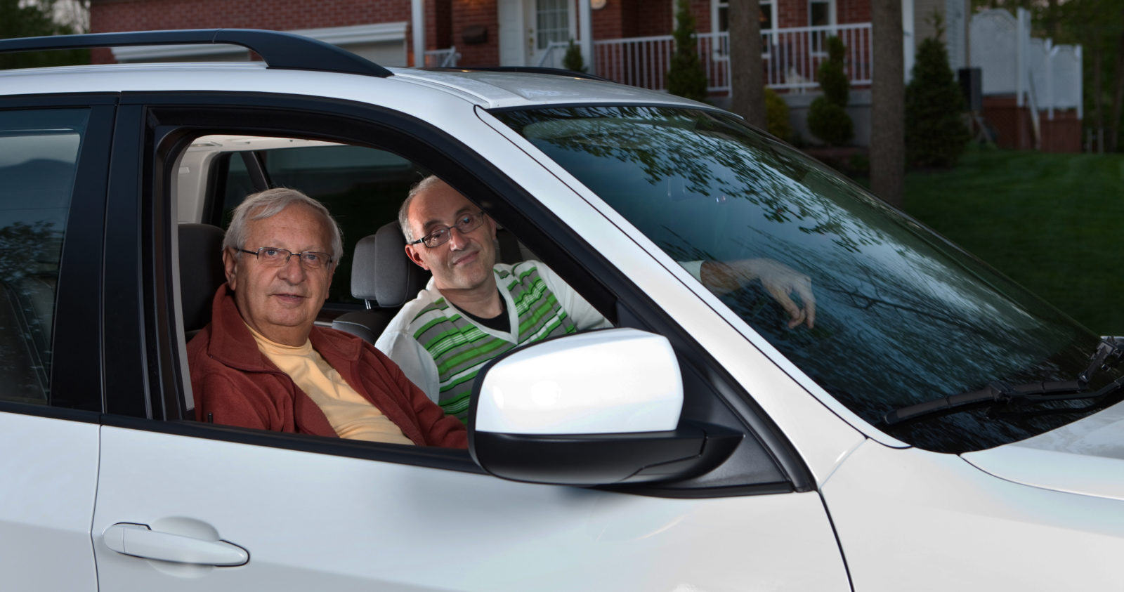 Two men parked in a car in front of house.