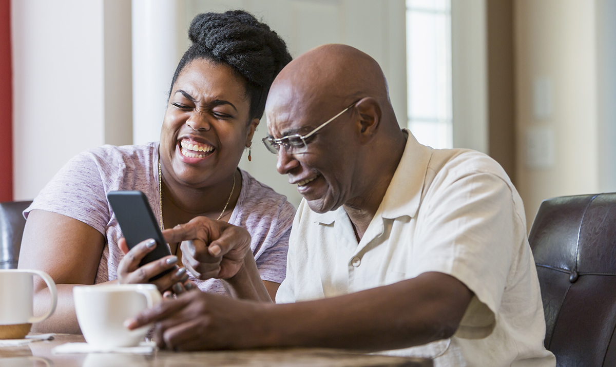 An older African-American man sits at home with his adult daughter, in her 30s at the dining room table, drinking coffee. The woman is showing her father something on her mobile phone and they are laughing.