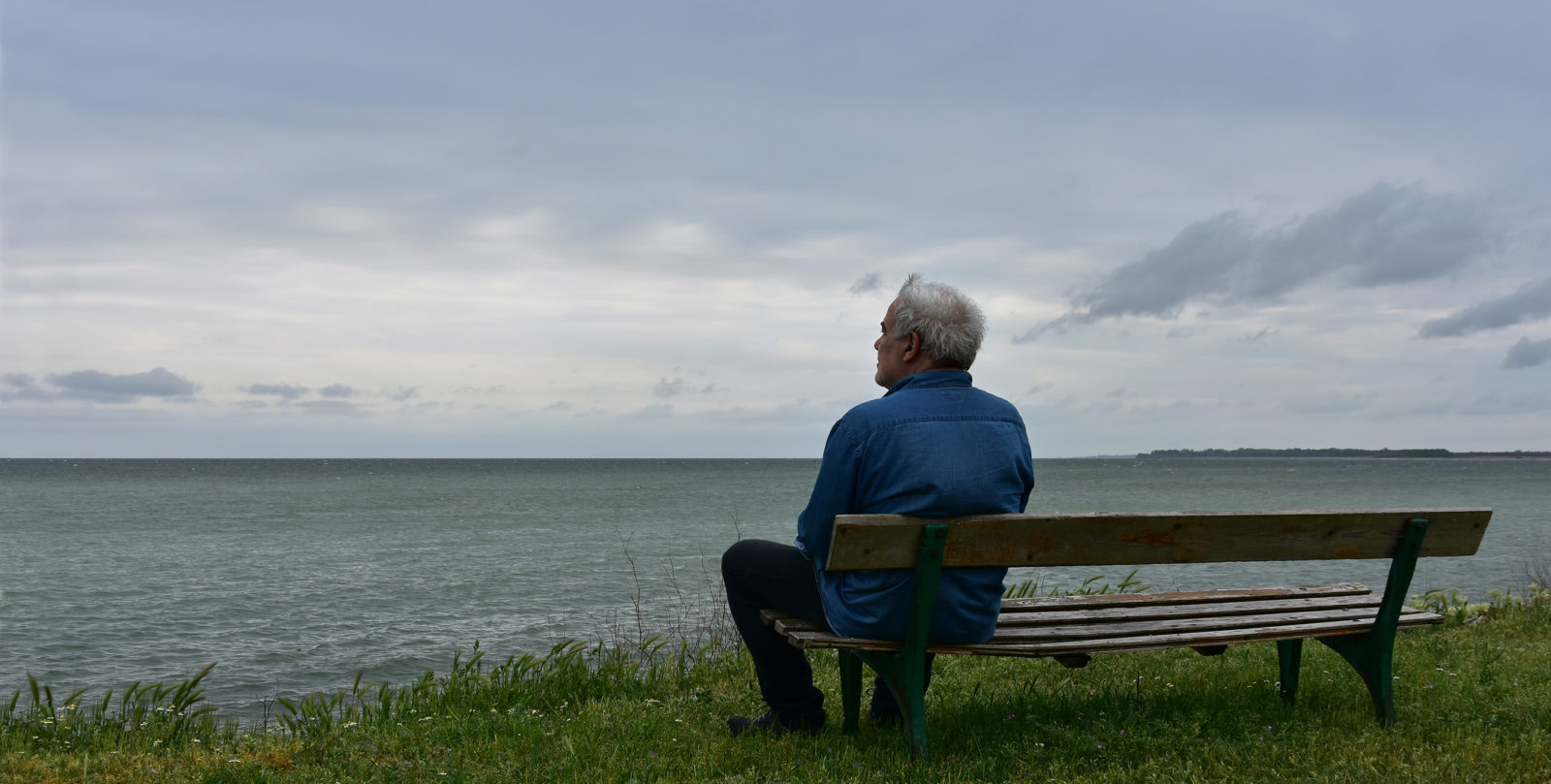 Elderly man sitting on an old wooden bench above the see in a cloudy day, contemplating the nostalgic sea-view