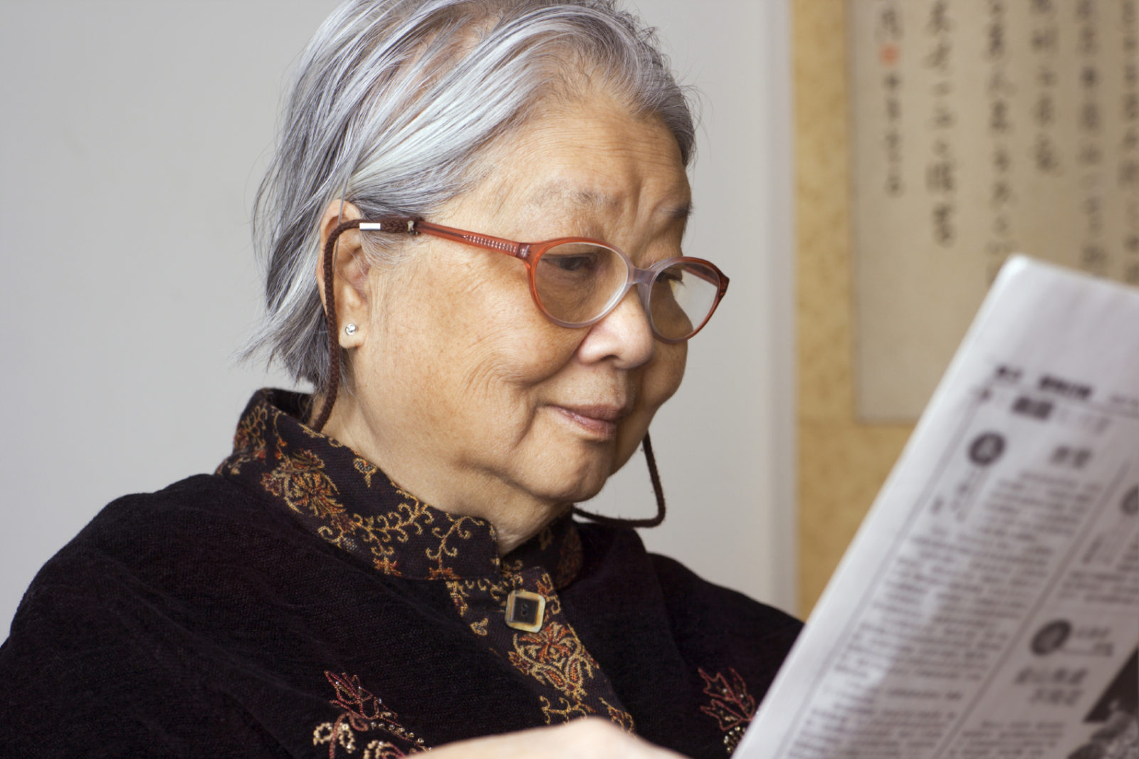 older Asian woman with slight smile and reading glasses, reading a newspaper