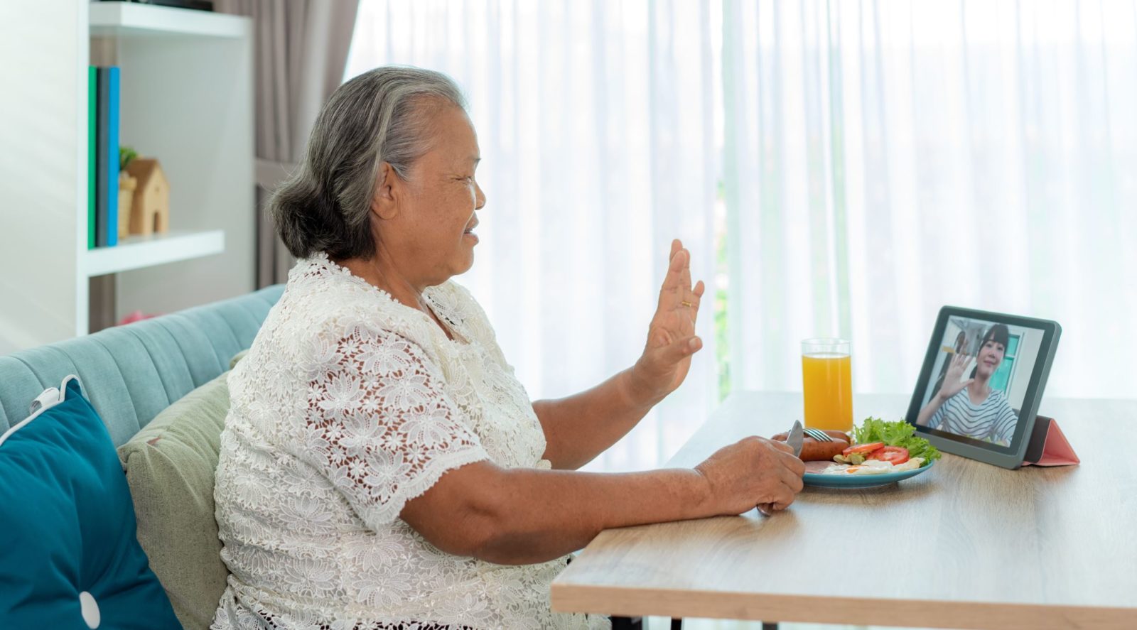 Older woman sits at a breakfast table and waves to someone on her laptop screen