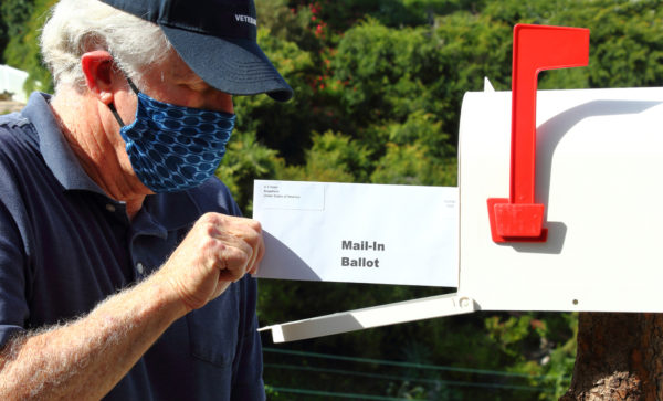 older man wearing a COVID face mask opens his mailbox and removes mail marked "mail-in ballot"