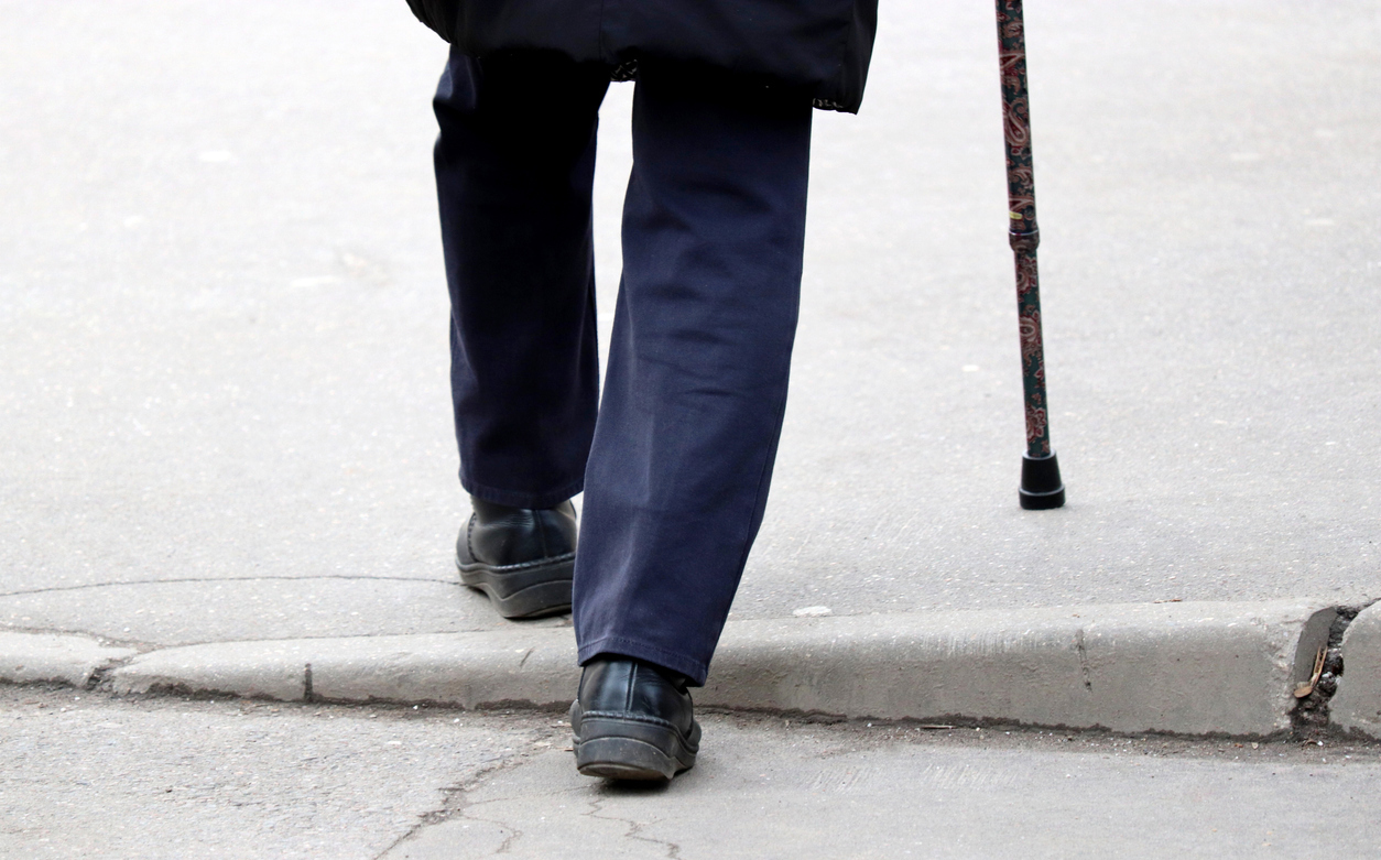 legs of an older person wearing an overcoat, dark slacks, and sturdy shoes, using a cane, steps up over an uneven sidewalk curb