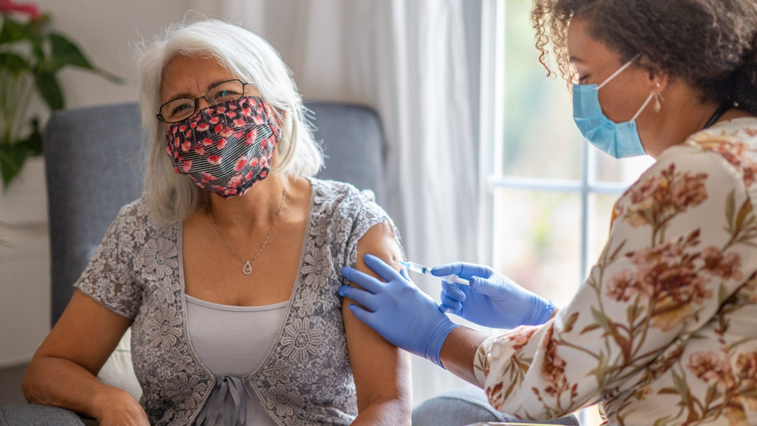 public health worker administers a vaccine in the upper arm of an older woman wearing a facemask