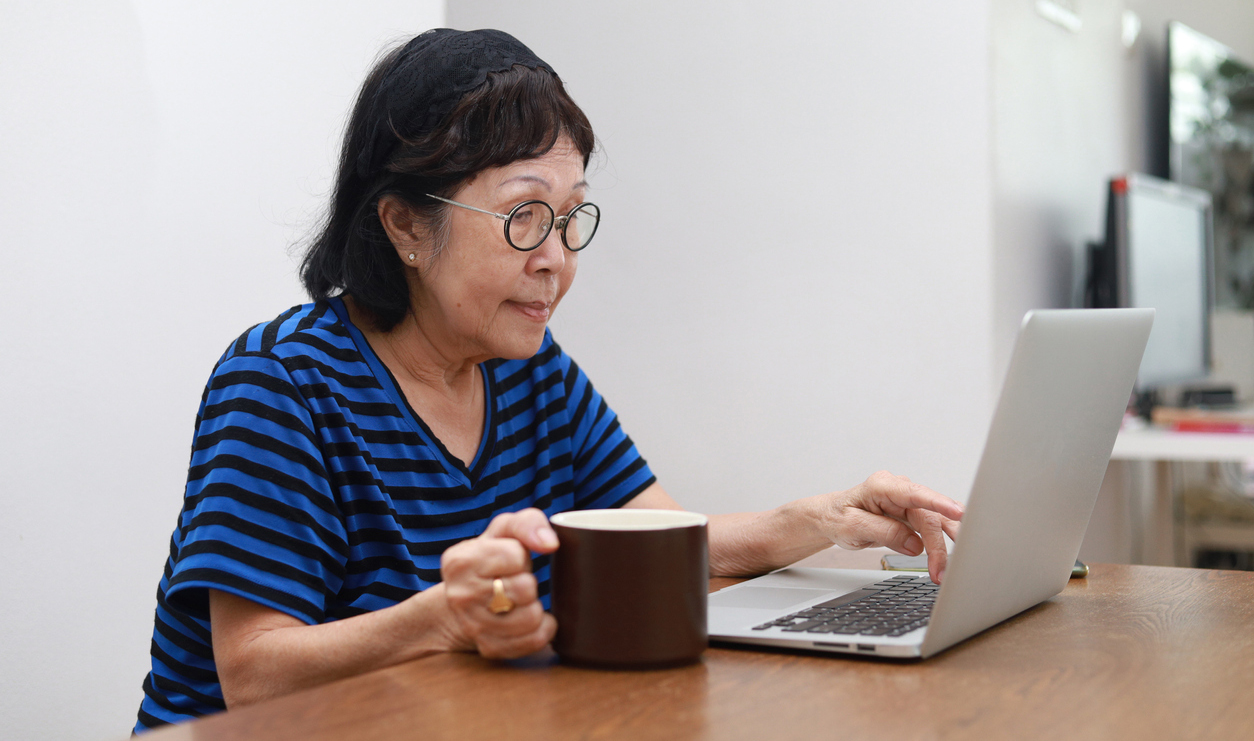 photo of an older woman of Asian descent working at a laptop computer with one hand on her coffee cup