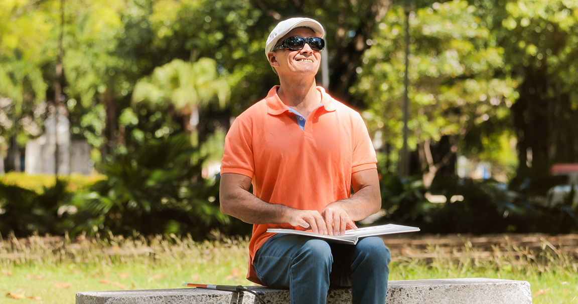 Latino man wearing sunglasses and reading Braille on a park bench