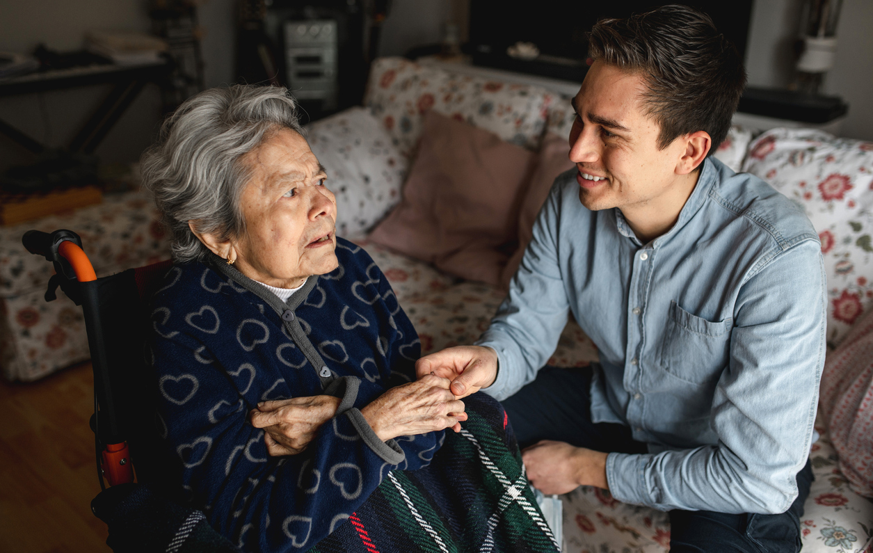 Young man sitting next to an older woman in wheelchair taking her hands while talking and smiling.