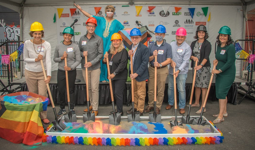 row of people at the Pride Place groundbreaking, wearing colorful hard hats and shoveling rainbow-colored sand