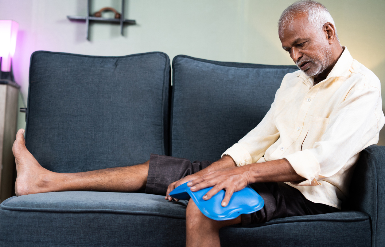 an older man on a couch uses a hot water bottle or ice pack on his knee