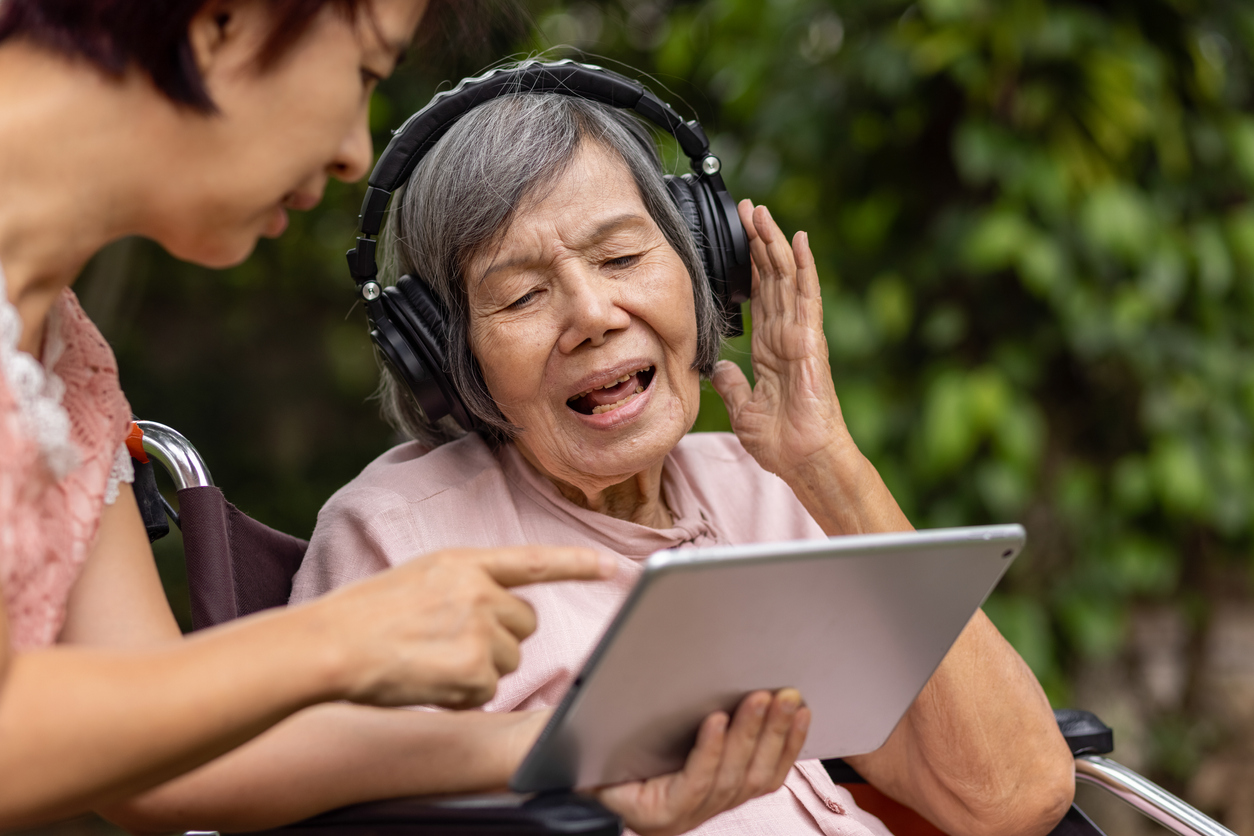 2 women, one younger pointing to an electronic tablet, an older woman sitting in a wheelchair wearing headphones reacts