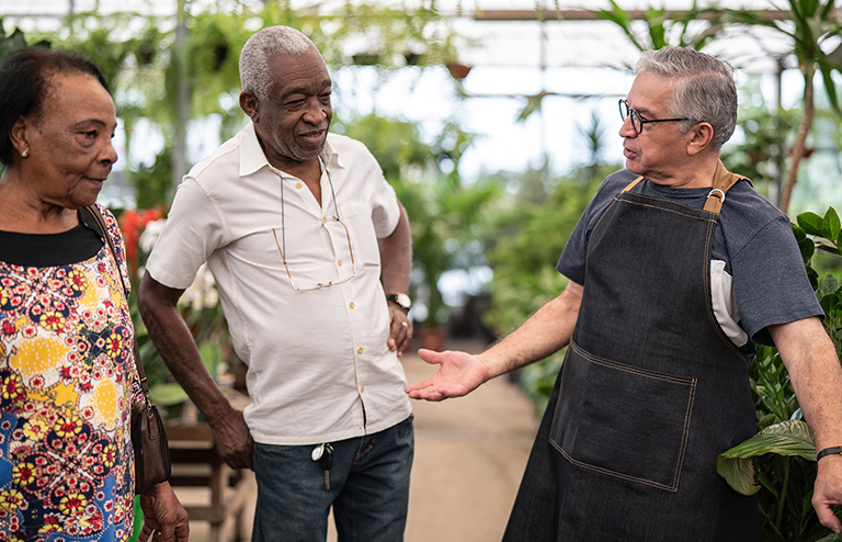 an older man working in a greenhouse talks with two older customers
