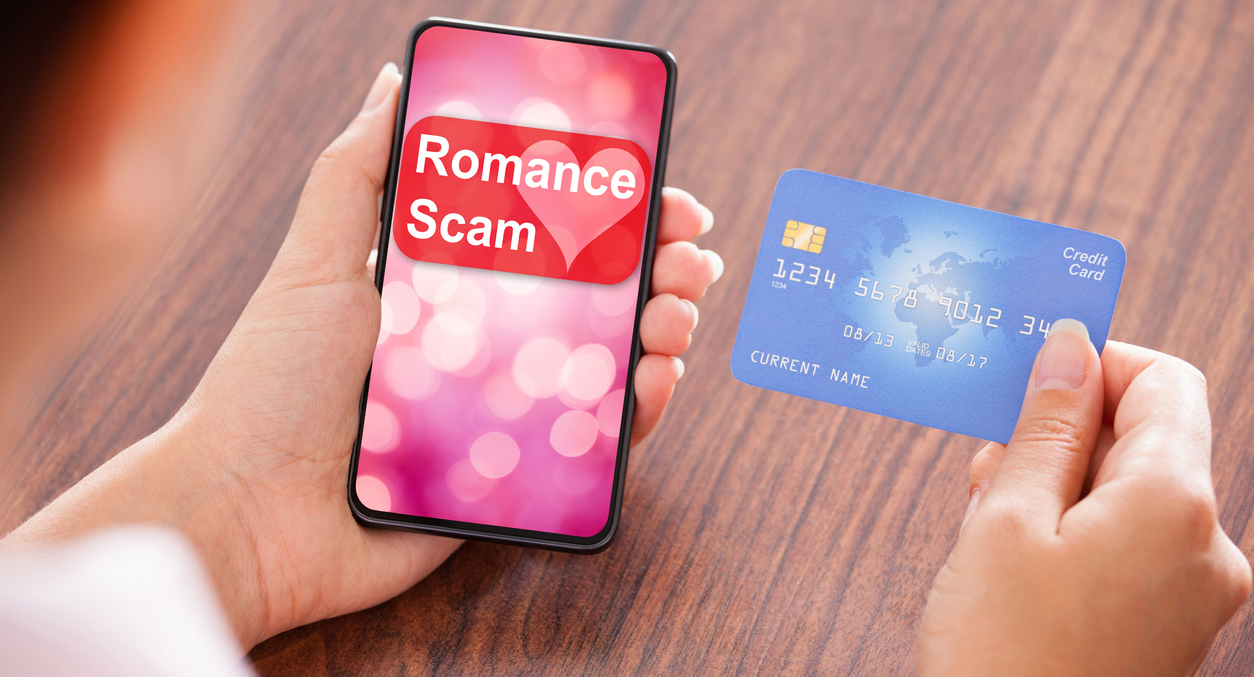 closeup of cell phone and nearby credit card - screen reads "Romance Scam"
