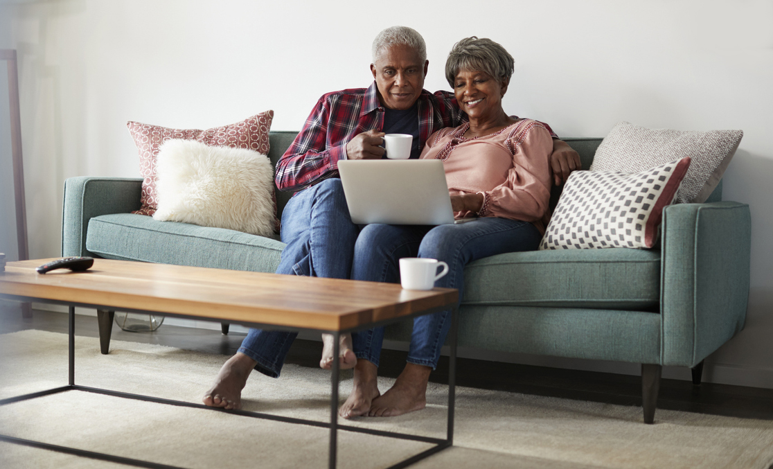 older man and woman sit on a living room couch and look at a laptop computer