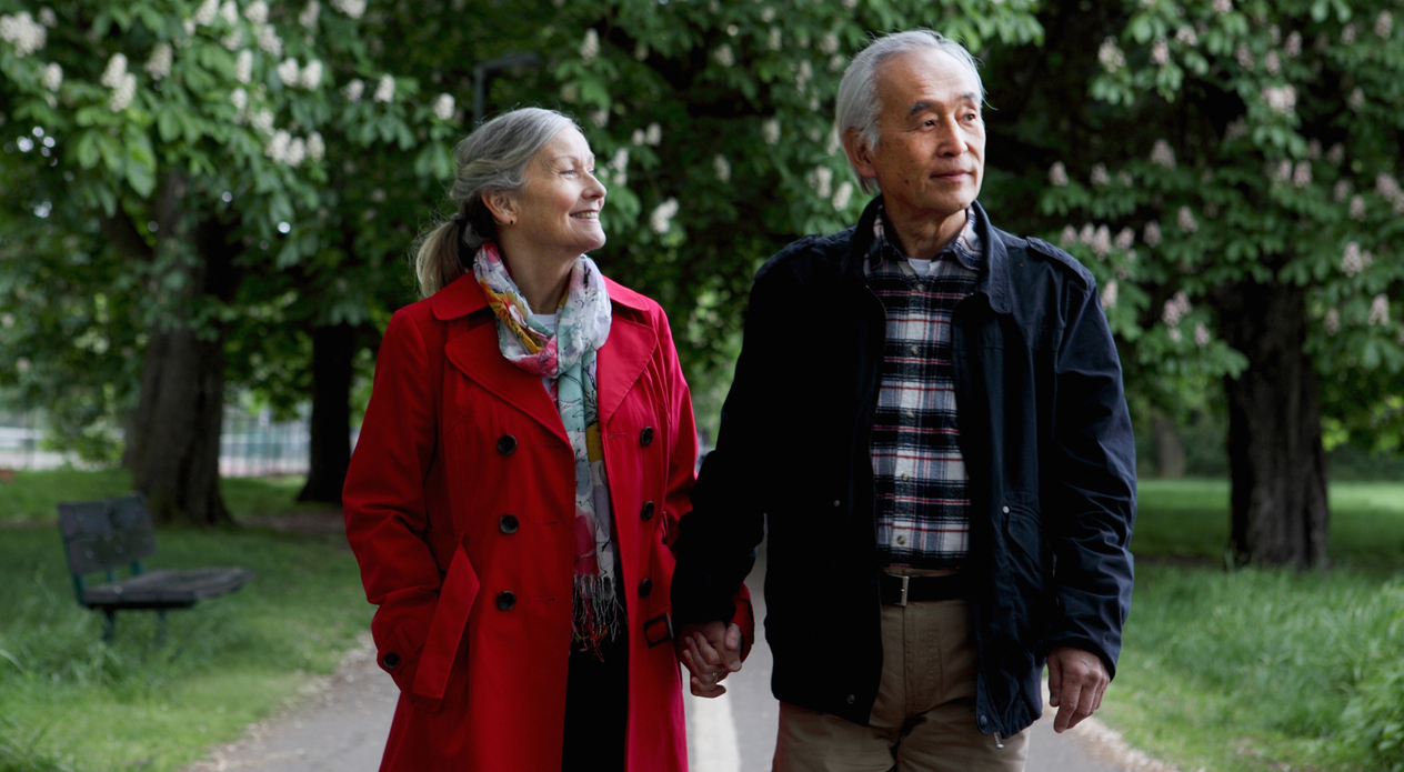 an older couple holding hands as they stroll through a park with green grass, trees, and a bench