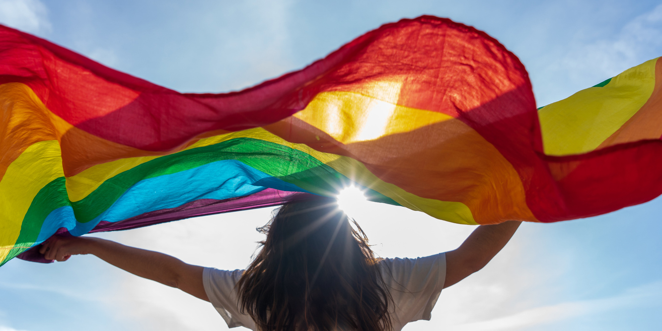 back of person with long hair, arms raised up, holding a rainbow striped scarf or flag in front of the sun