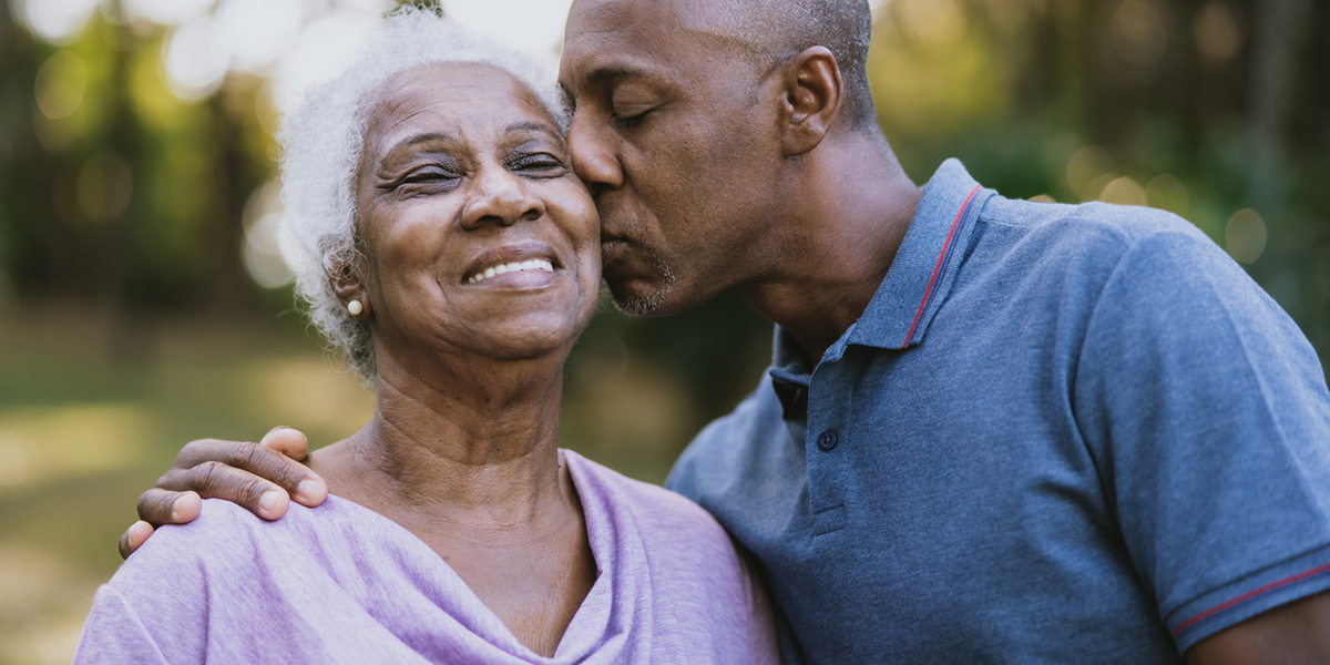 old African American couple outdoors, he leans down to kiss her cheek, she looks up and smiles